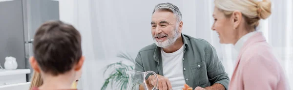 Älterer Mann lächelt neben verschwommenen Enkeln und Ehefrau in Küche, Banner — Stockfoto