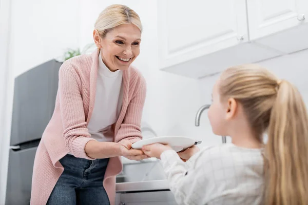 Blurred girl holding plate while helping happy granny in kitchen — Stock Photo