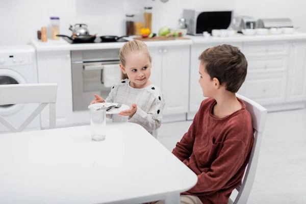 Улыбающаяся девушка держит тарелку со столовыми приборами рядом с братом, сидящим за кухонным столом — стоковое фото