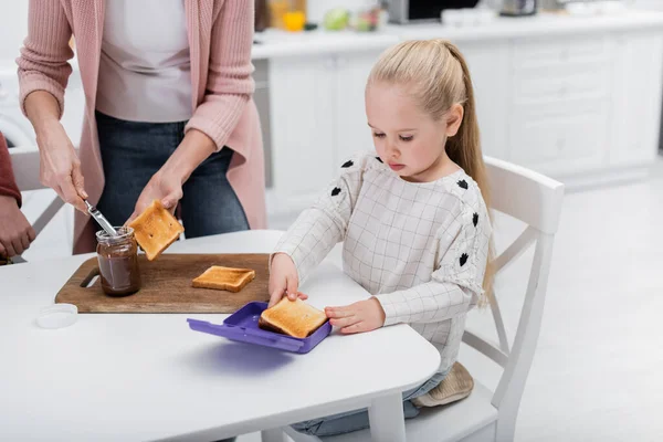 Frau bereitet Toast mit Schokoladenpaste in der Nähe der Enkelin mit Lunchbox in der Küche zu — Stockfoto