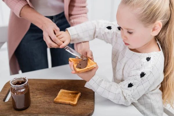 Kleines Mädchen verteilt Schokoladenpaste auf Brot, während es mit Oma Sandwiches zubereitet — Stockfoto