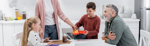 Senior man smiling near wife preparing sandwiches for grandchildren in kitchen, banner — Stock Photo