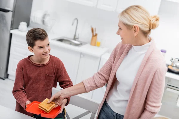 Smiling woman putting sandwich in lunch box of happy grandson in kitchen — Stock Photo