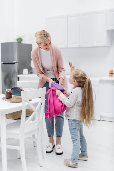 Smiling woman holding lunch box while packing backpack of granddaughter in kitchen — Stock Photo