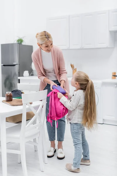 Femme souriante tenant boîte à lunch tout en emballant sac à dos de petite-fille dans la cuisine — Photo de stock