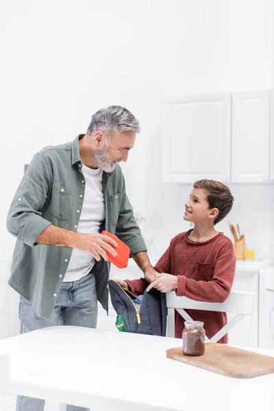 Hombre de pelo gris sosteniendo la lonchera mientras empaca la mochila de su nieto sonriente - foto de stock