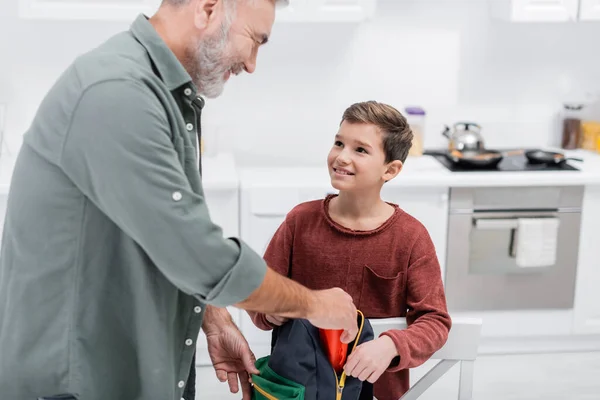 Lächelnder Senior packt Rucksack seines Enkels in Küche — Stockfoto