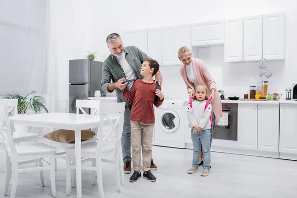 Smiling grandparents putting backpacks on kids in kitchen — Stock Photo