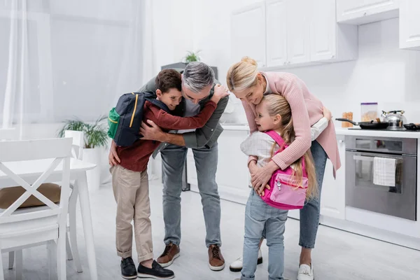 Abuelos felices abrazando a los niños con mochilas en la cocina - foto de stock