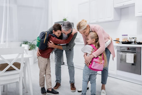 Happy grandparents embracing children with backpacks in kitchen — Stock Photo