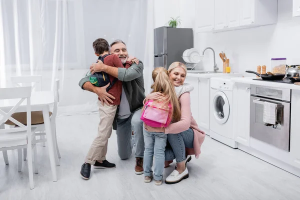 Smiling couple hugging grandchildren with backpacks in kitchen — Stock Photo