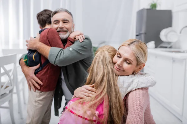 Happy couple embracing grandchildren with backpacks in kitchen — Stock Photo