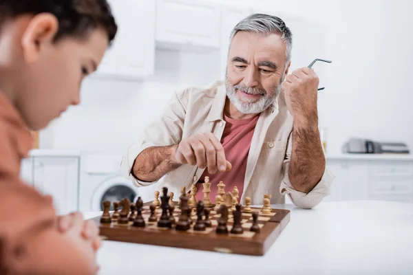 Pleased senior man playing chess with blurred grandson in kitchen — Stock Photo