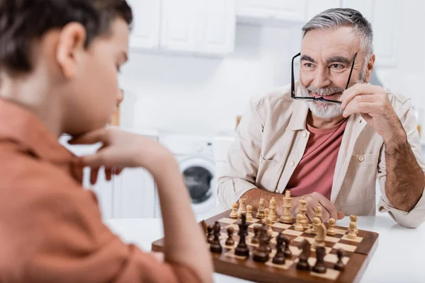Happy senior man holding eyeglasses while playing chess with blurred grandson — Stock Photo