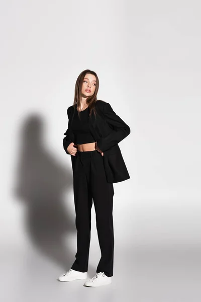 Full length view of brunette woman in black suit and white sneakers on grey with shadow — Stock Photo