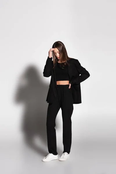 Vista integrale della donna in completo nero e sneakers bianche in piedi con mano sul fianco su grigio con ombra — Foto stock