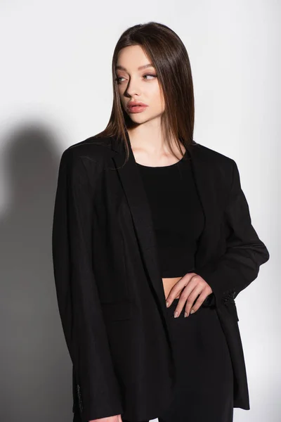 Stylische Frau in schwarzer Kleidung, die mit der Hand auf der Taille steht und wegschaut auf grau mit Schatten — Stockfoto