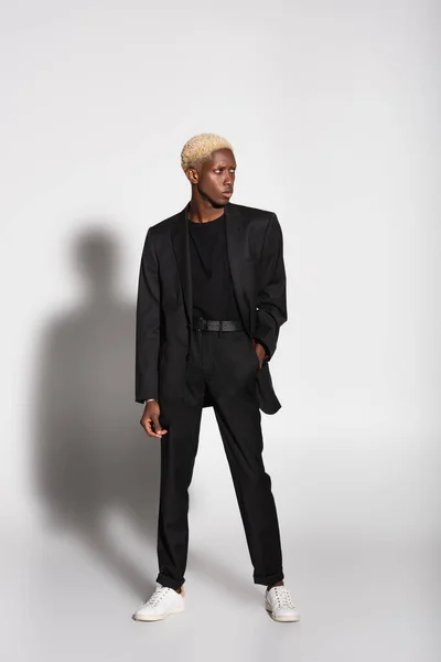Vista integrale dell'uomo afroamericano in completo nero e sneakers bianche in piedi con mano in tasca su grigio con ombra — Foto stock
