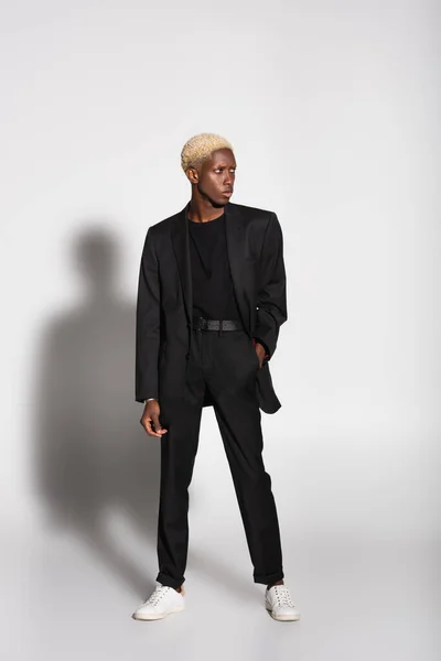 Full length view of african american man in black suit and white sneakers standing with hand in pocket on grey with shadow — Stock Photo