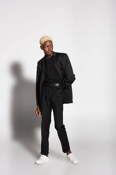 Biondo afroamericano in abito nero e sneakers bianche in posa su grigio con ombra — Foto stock