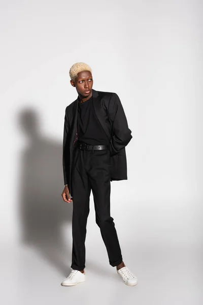 Blonde african american man in black suit and white sneakers posing on grey with shadow — Stock Photo