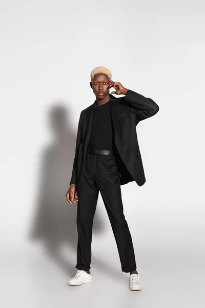 Blonde afrikanisch-amerikanische Mann im schwarzen Anzug posiert mit der Hand nahe Gesicht auf grau mit Schatten — Stockfoto