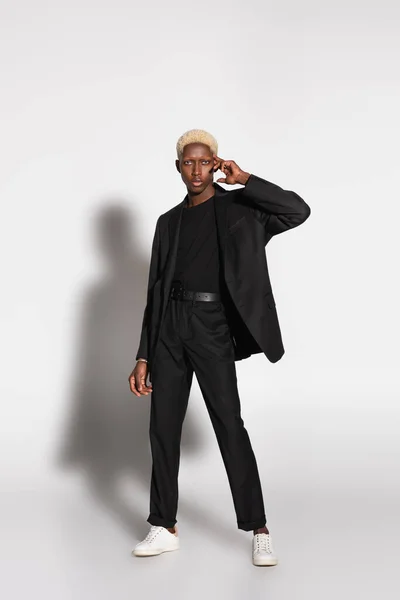 Blonde african american man in black suit posing with hand near face on grey with shadow — Stock Photo