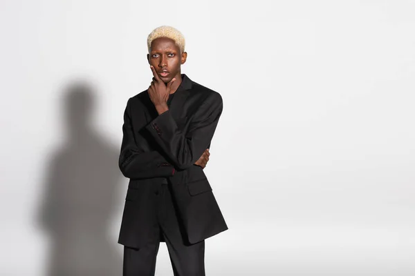 Blonder afrikanisch-amerikanischer Mann im schwarzen Anzug hält die Hand in der Nähe des Gesichts, während er auf grau mit Schatten posiert — Stockfoto