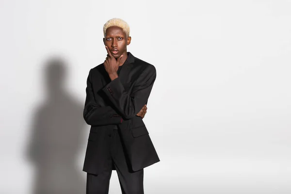 Blonde african american man in black suit holding hand near face while posing on grey with shadow — Stock Photo