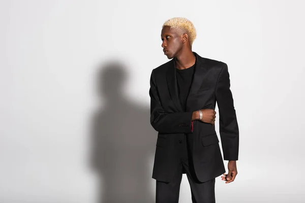 Blonde african american man in black suit and silver bracelet looking away on grey with shadow — Stock Photo