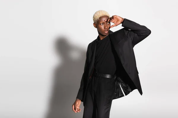 Homme afro-américain à la mode avec des cheveux blonds et la main près du visage regardant loin sur gris avec ombre — Photo de stock