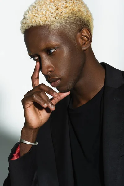 Jeune homme afro-américain aux cheveux blonds posant avec la main près du visage sur gris — Photo de stock