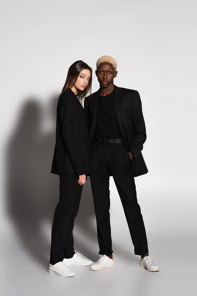 Full length view of blonde african american man and brunette woman in black suits on grey with shadow — Stock Photo