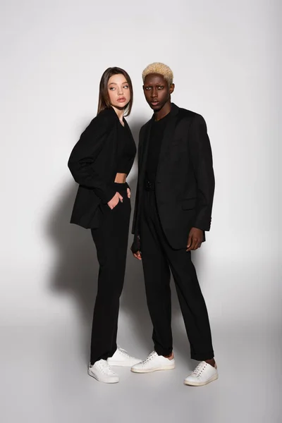 Full length view of brunette woman with hands in pockets and african american man with bleached hair on grey with shadow — Stock Photo