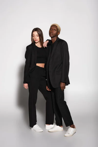 Pareja multiétnica de moda en ropa negra y zapatillas blancas posando en gris con sombra - foto de stock