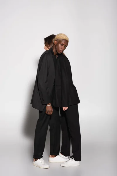 Vue pleine longueur de couple interracial élégant en costumes noirs s'appuyant l'un sur l'autre sur gris avec ombre — Photo de stock