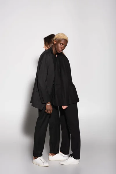 Full length view of stylish interracial couple in black suits leaning on each other on grey with shadow — Stock Photo