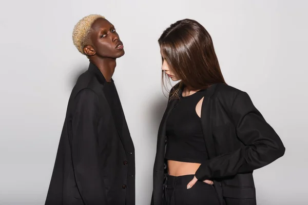 Brunette femme debout avec la main sur la hanche près blond afro-américain homme regardant caméra sur gris — Photo de stock