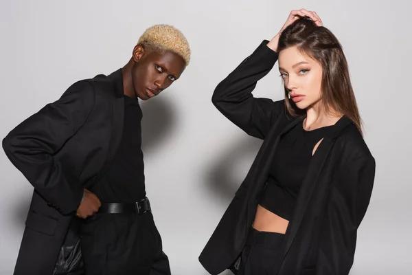 Young and stylish woman touching hair near blonde african american man posing with hand on waist on grey with shadow — Stock Photo