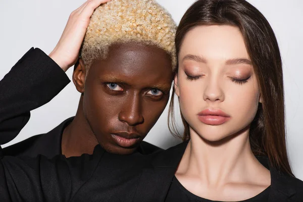 Jolie femme brune aux yeux fermés touchant les cheveux d'homme blond afro-américain sur gris — Photo de stock