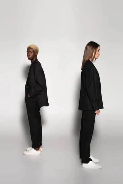 Homme afro-américain avec les cheveux blanchis debout avec la main dans la poche près de femme élégante sur gris avec ombre — Photo de stock