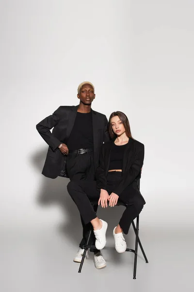 Homme afro-américain à la mode avec la main sur la taille près de la femme sur chaise sur gris avec ombre — Photo de stock