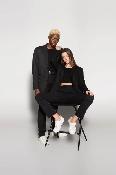 Woman in black suit and white sneakers sitting on chair near blonde african american man on grey with shadow — Stock Photo