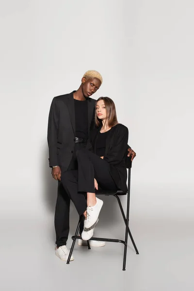 Blonde african american man looking at camera near brunette woman on chair on grey with shadow — Stock Photo