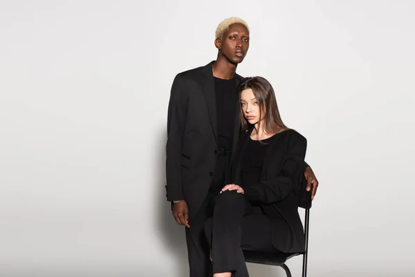 Blonde african american man looking at camera near brunette woman on chair on grey — Stock Photo