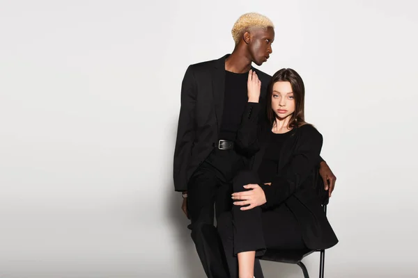 Pretty woman in white clothes sitting on chair near blonde african american man on grey — Stock Photo