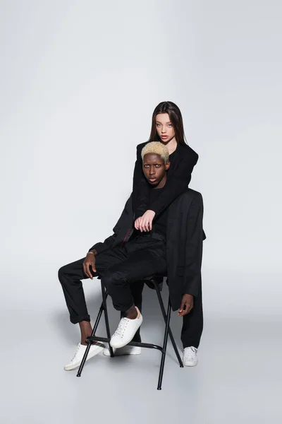 Brunette woman hugging african american man on chair on grey with shadow — Stock Photo