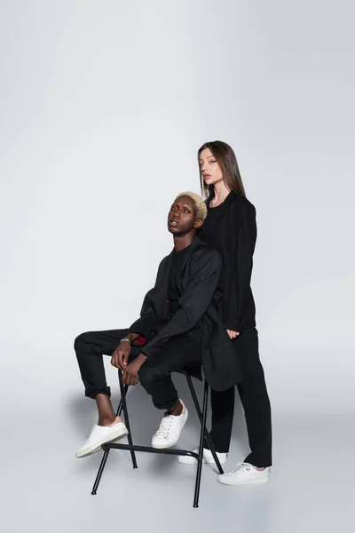 African ameican man with bleached hair sitting on chair near trendy brunette woman on grey with shadow — Stock Photo