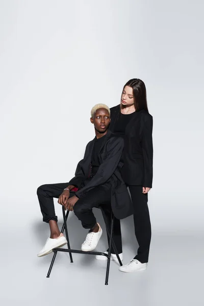 Woman in black suit looking at blonde african american woman sitting on chair on grey with shadow — Stock Photo