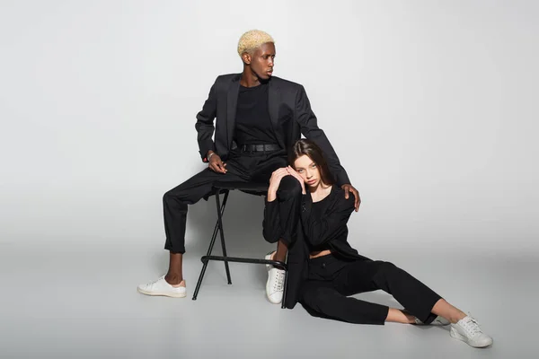 Stylish woman looking at camera while sitting on floor near blonde african american man on chair on grey with shadow — Stock Photo